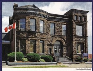 Large sandstone two story building with large windows, entrance with stairs and Canada flag on pole at left