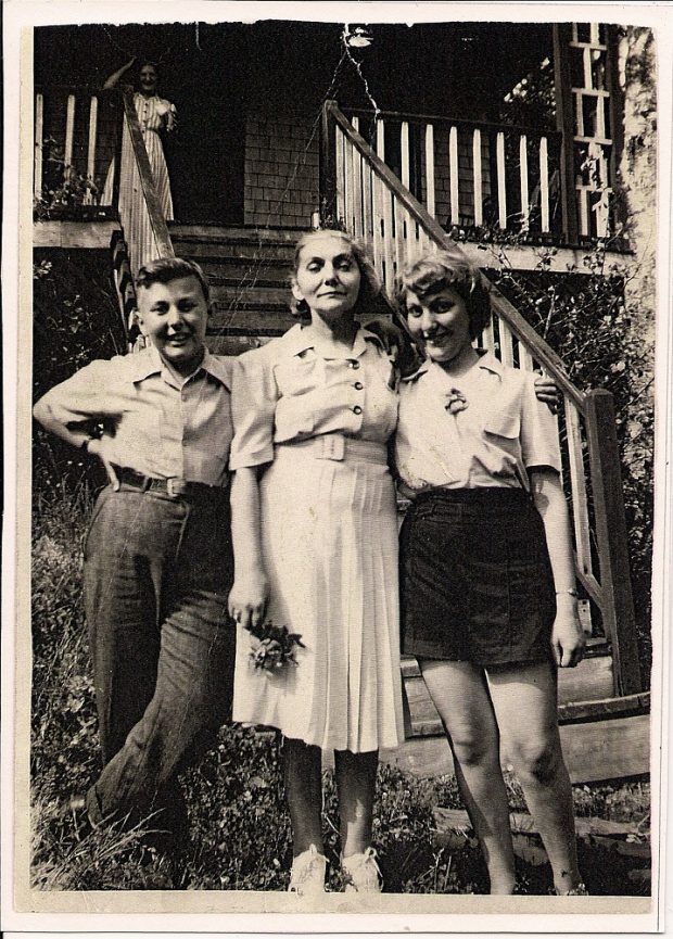 Woman standing at bottom of wooden steps leading to cottage with teenaged daughter on her left arm and teenaged son at her right arm