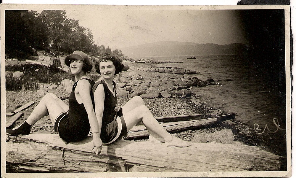 Two women in bathing suits seated on a large piece of driftwood on a beach with their backs together