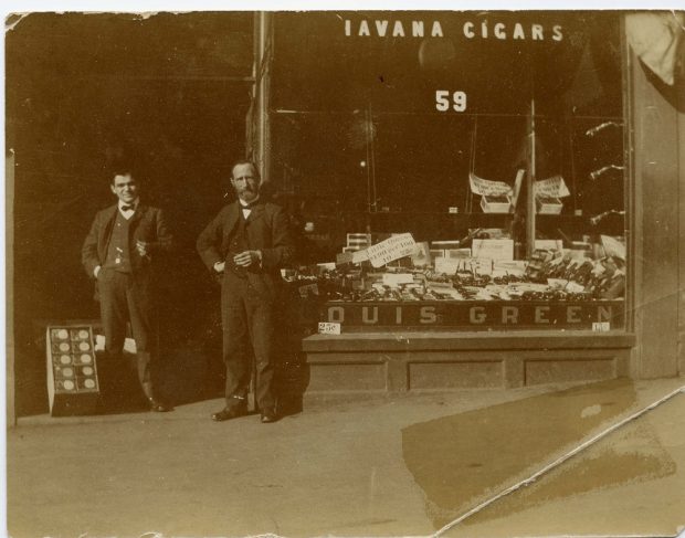 Storefront with large glass window with “Havana Cigars” written at top of window and “Louis Green” printed at bottom – the window displays cigars and tobacco products – two men standing in doorway