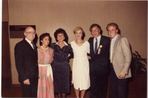 Three men and three women – men in business suits, women in dresses – standing with arms linked