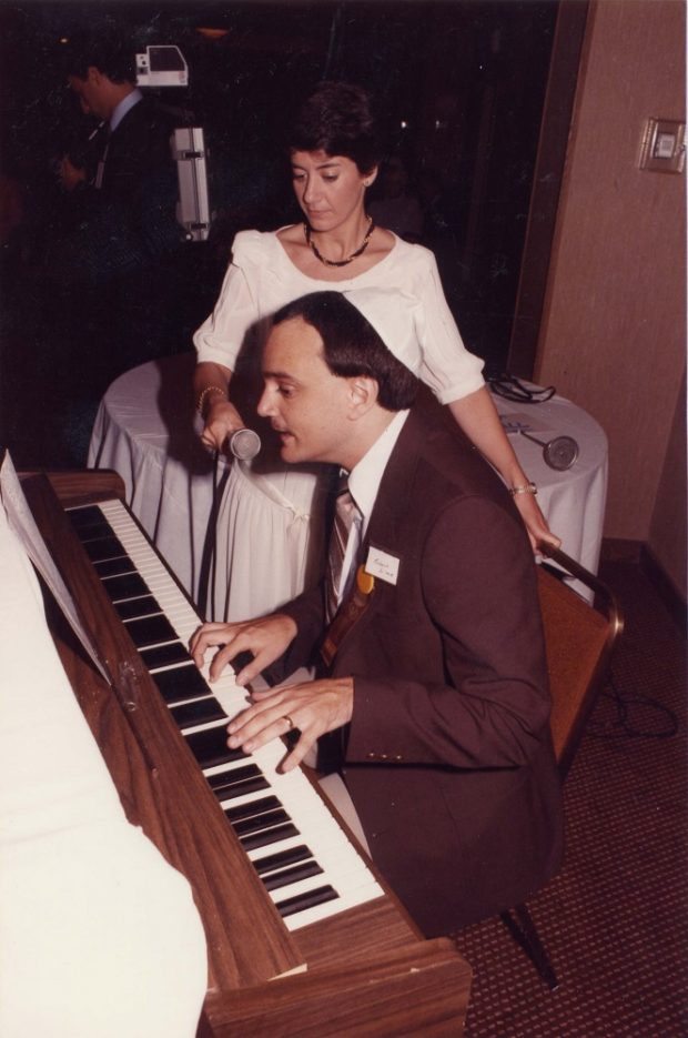 Man seated at piano with woman holding a microphone to his mouth