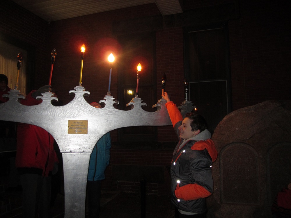 Large grey painted Chanukah menorah with some candles lit and teenaged boy reaching up to adjust lights