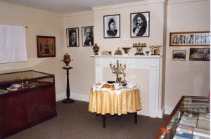 Salle d’exposition du Musée où l’on voit deux présentoirs vitrés, une table préparée pour un repas du Shabbat, un assortiment de menorahs de Hanoukka en laiton sur le manteau de cheminée et des photographies sur le mur.