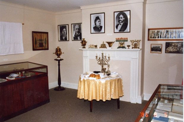 Exhibition room in Museum with two display cabinets, table set for Sabbath meal, assortment of brass Chanukah menorahs on mantelpiece and photographs on wall