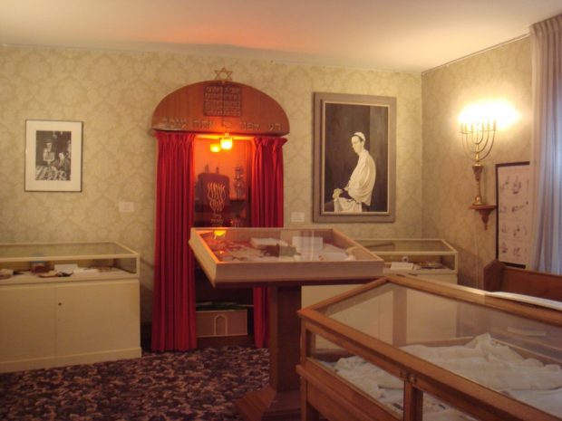Museum exhibition with three display cabinets and wooden Holy Ark and painted portrait of man in prayer by Fred Ross at right