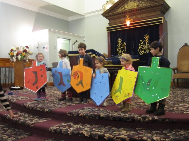 Six children standing on platform at front of Synagogue – each child holding a life-size colourful cardboard dreidl
