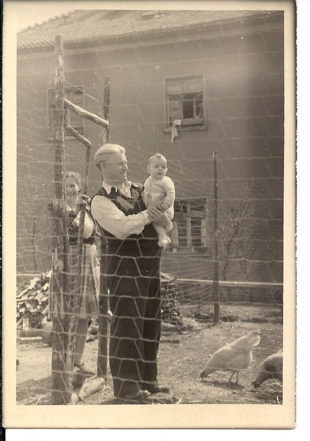 Man with white hair holding infant son in his arms and wife in background – all are posing behind a barbed wire fence