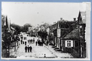 Rue de village non asphaltée bordée de chaque côté de petits bâtiments en bois construits sur deux niveaux. Plusieurs hommes y déambulent ou forment des groupes.