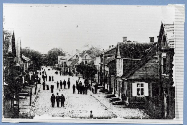 Unpaved village street with small two story wooden buildings on either side and several men walking or gathered in street