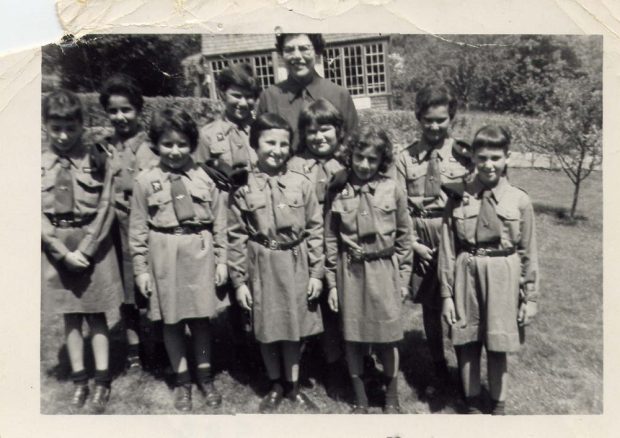 9 girls in Brownie uniform dresses with leader in Brownie uniform standing behind