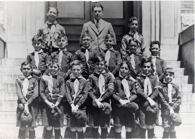 14 boys in Cub uniforms with hats in hand standing in two rows on stairs with leaders in third row behind