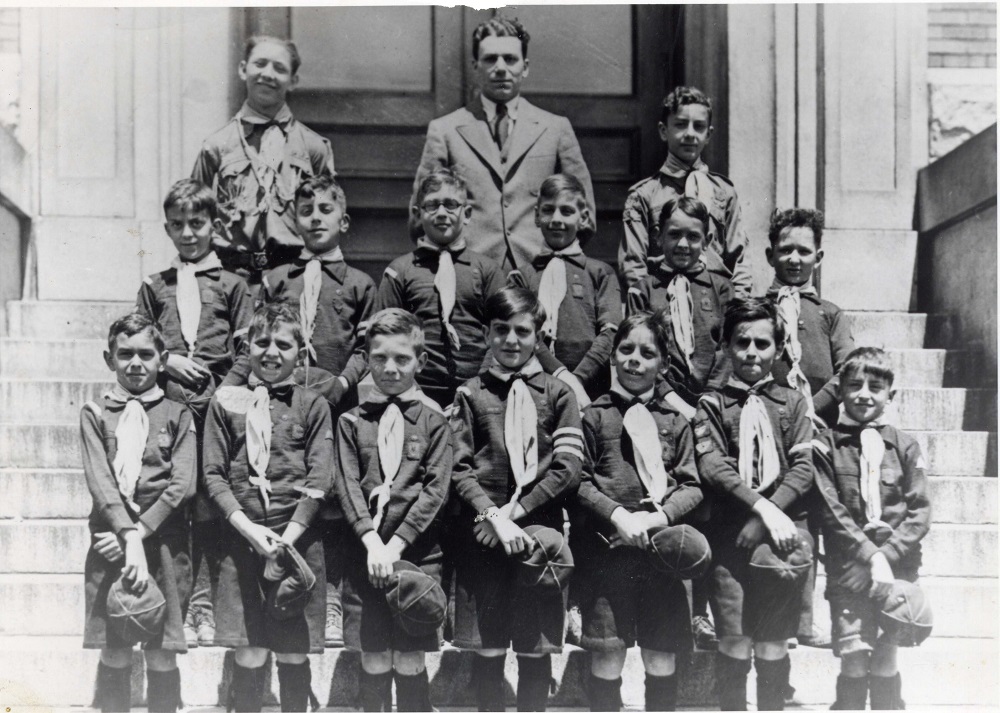 14 boys in Cub uniforms with hats in hand standing in two rows on stairs with leaders in third row behind