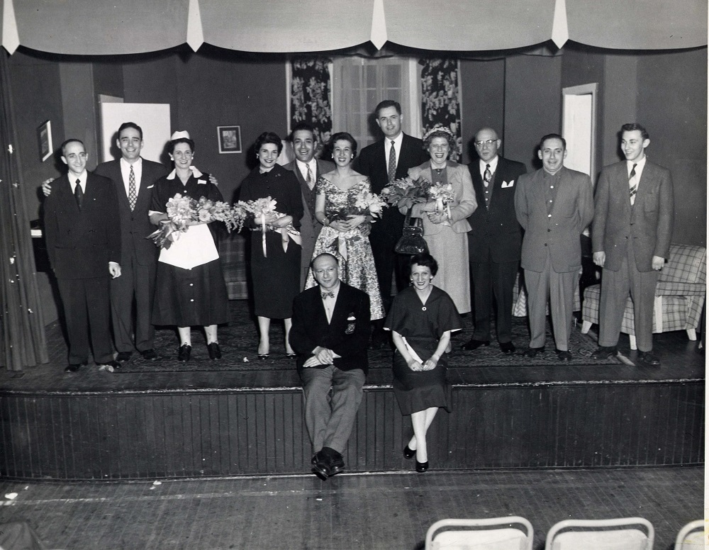 11 men and women in costumes standing at front of stage with man and woman seated on stage edge in front of them