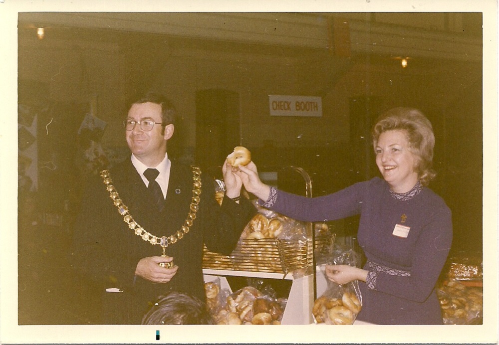 Colour photograph showing man in business with mayoral chain of office and woman standing in front of baking table at Hadassah Bazaar