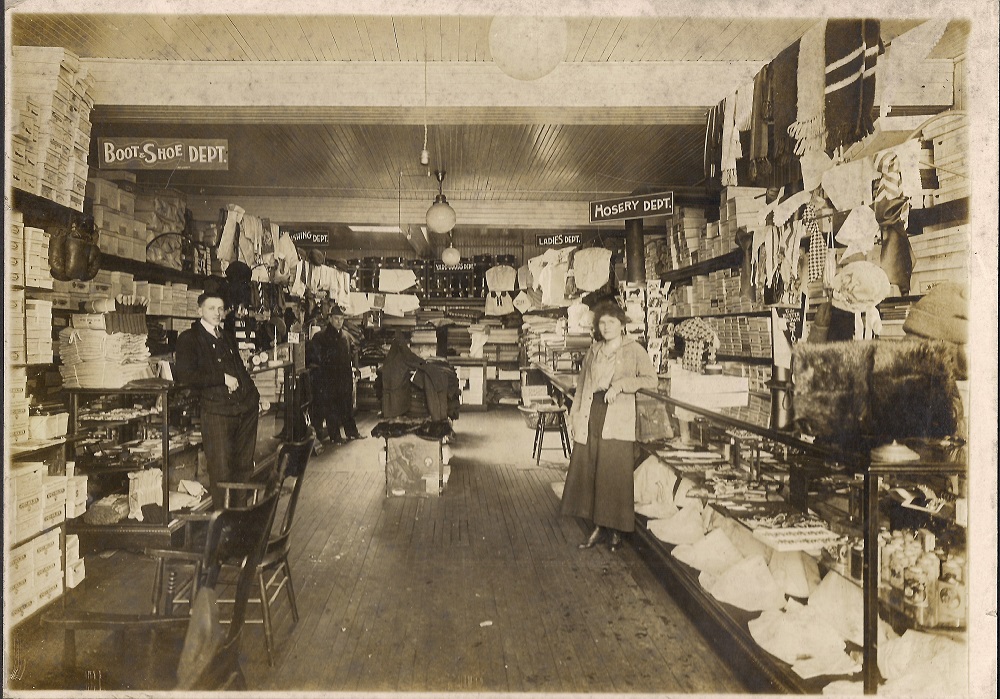 View inside a narrow dry goods shop with counters and shelves with boxes on either side and at the back, man at left, woman at right