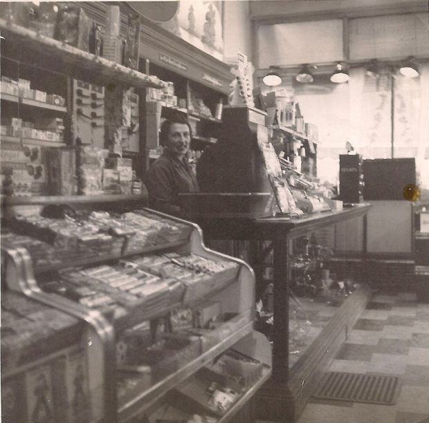 View shows interior of store with counter hilding small items, shelves with boxes and young woman at counter beside large cash register