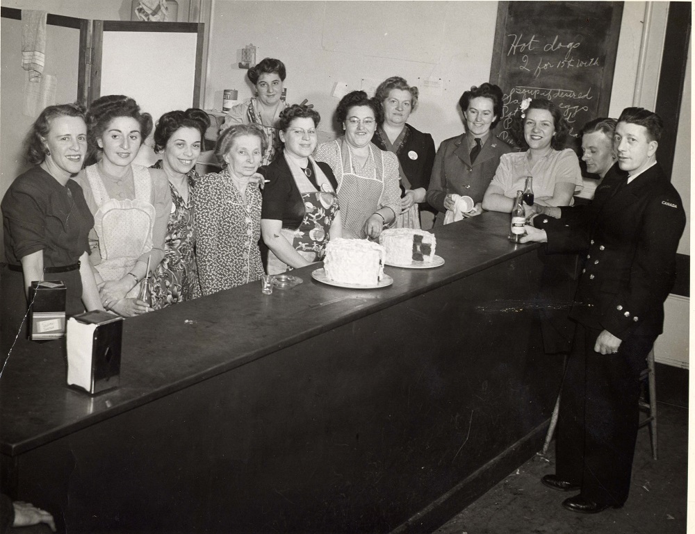 10 women lined up behind long counter, some wearing aprons – two men in naval uniforms at far end