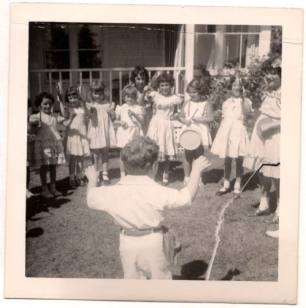 Boy standing with back to camera directing group of girls wearing summer dresses and holding assorted instruments including triangles and tambourines