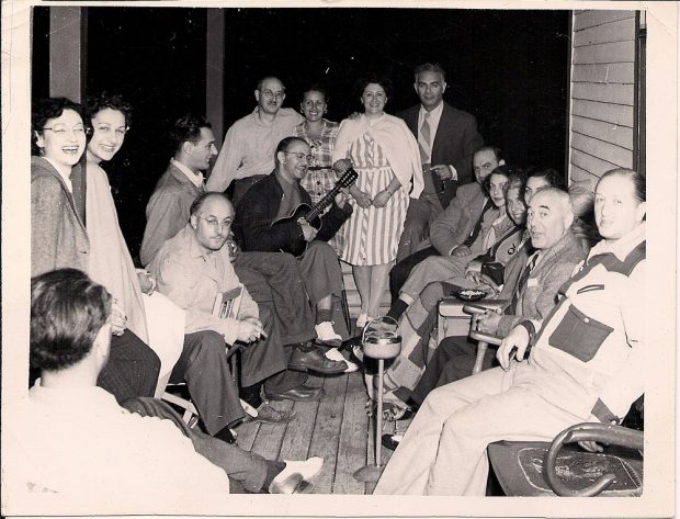 16 men and women seated on a wooden verandah – one man holding a ukelele