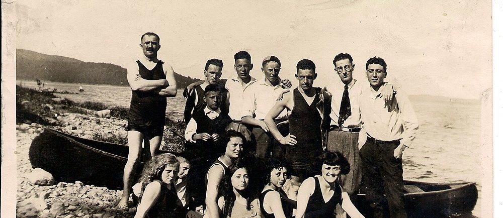 13 friends – some in bathing suits, others in pants and shirts gathered in front of a beached canoe