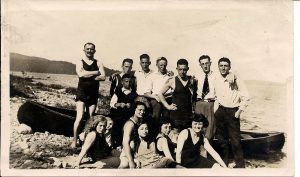 13 friends – some in bathing suits, others in pants and shirts gathered in front of a beached canoe