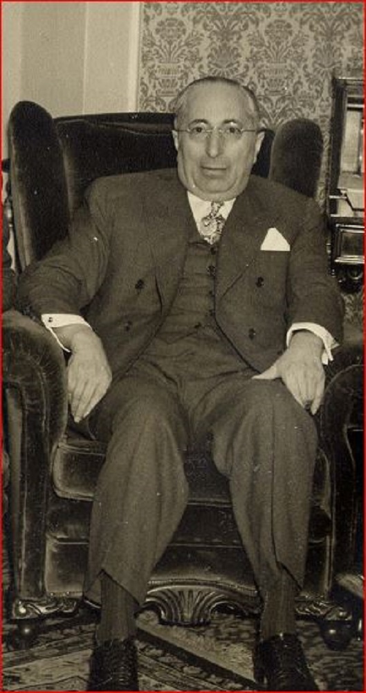 Man wearing a three piece suit and glasses seated in a wingback chair