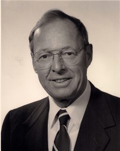 Portrait officiel d’un homme d’âge moyen portant complet et lunettes.