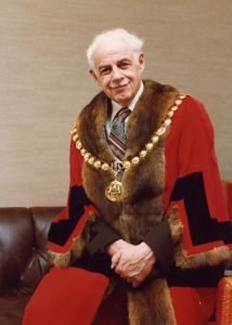 Portrait officiel d’un homme aux cheveux gris portant l’uniforme protocolaire du maire, y compris la chaîne. Il s’agit en fait d’une toge de magistrat, un vêtement ample et long de couleur rouge garnie de fourrure brune autour du col et le long de l’ouverture et au bas des manches, également amples.