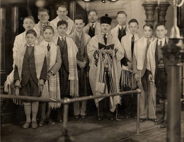 13 boys and men in suits and with prayer shawls over their shoulders with the Rabbi holding an opened prayer book
