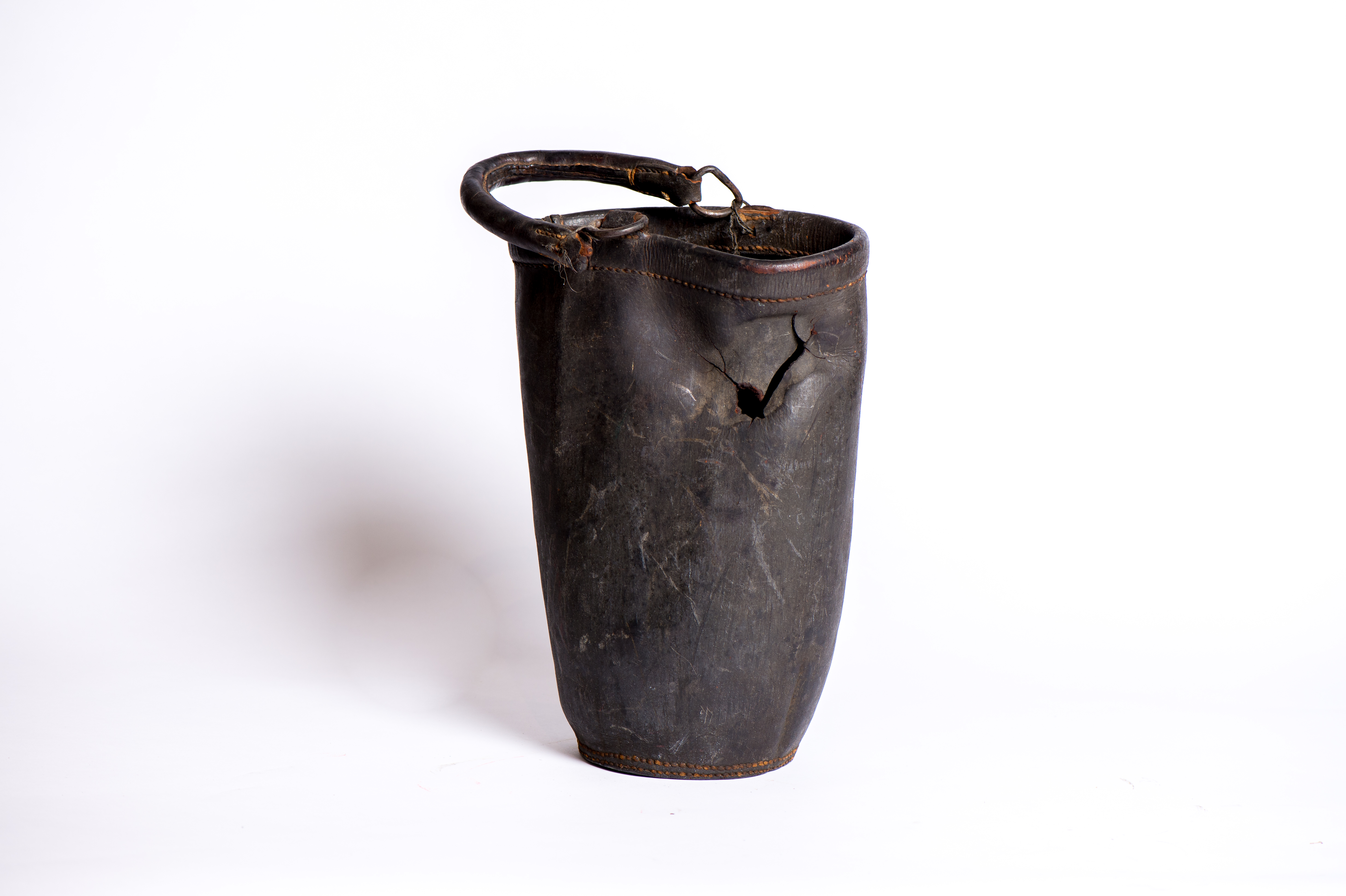 Photograph of an oval, black leather fire bucket on a white background. The bucket has a crack in it, clearly showing that it is old and worn.