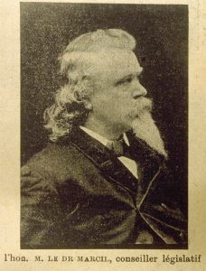 Photographie en noir et blanc d’un homme de profil portant un veston élégant noir. Sa chevelure frisée dépasse son cou, tandis qu’une moustache massive ainsi qu’une longue barbiche couvrent sa bouche et son menton.