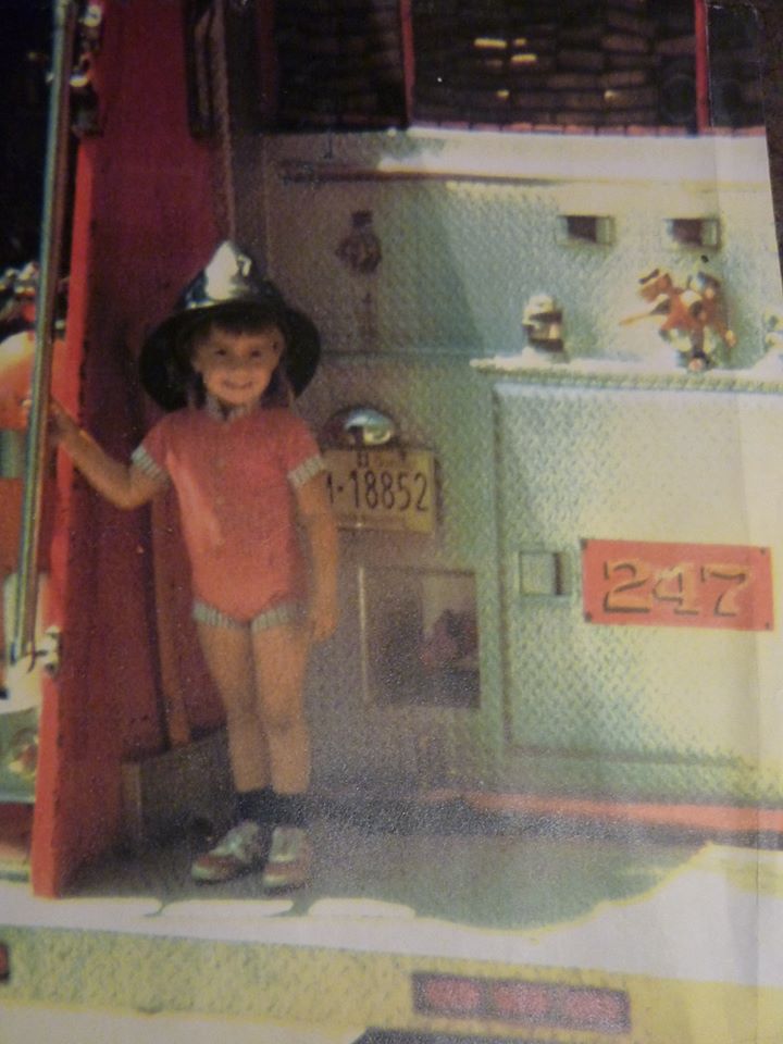Colour photograph of a four-year-old girl standing at the back of a fire truck, smiling and wearing a firefighter’s helmet.