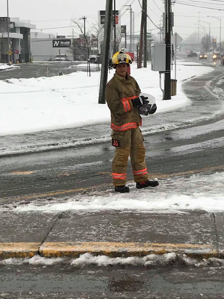 Photograph of a firefighter in his firefighting gear; he is at an intersection, holding a bucket to collect donations from motorists.
