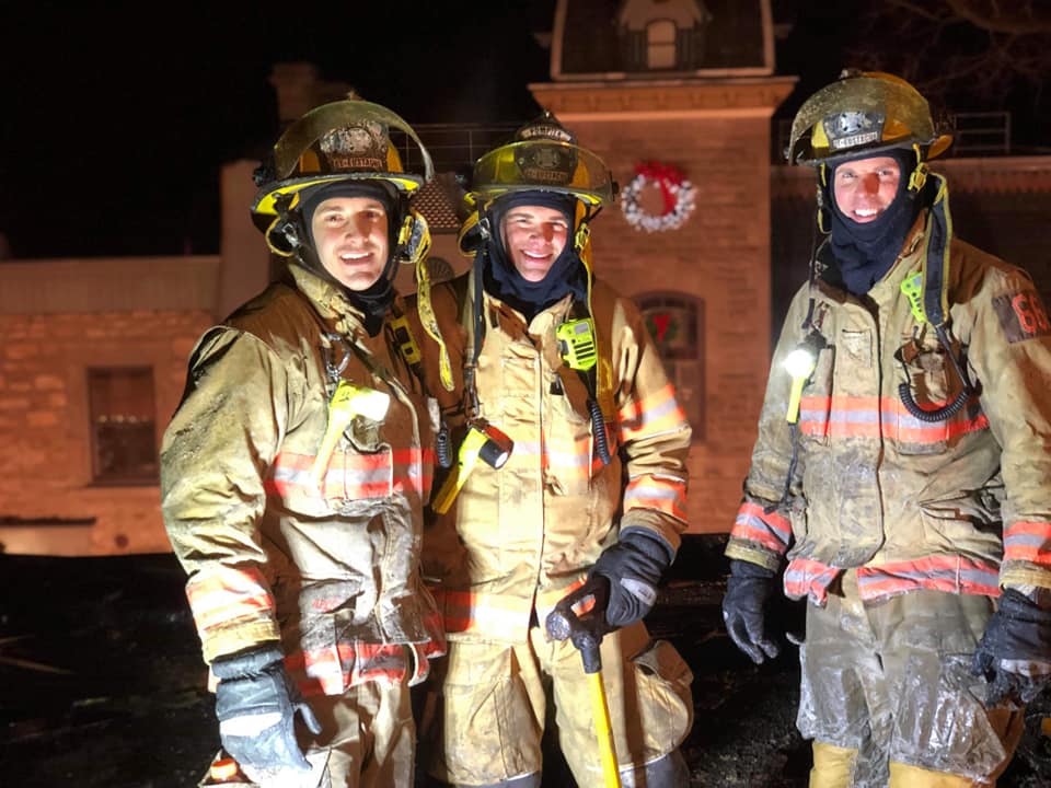 Photograph of three smiling firefighters in firefighting gear, in front of a church, at night, under the lights.