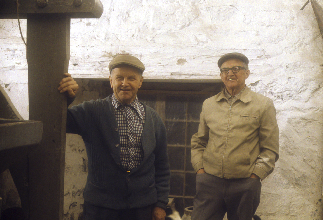 Photograph of two men in their seventies, smiling broadly, in front of a white concrete wall. The man on the left is wearing a check shirt under a navy cardigan. The man on the right has his hands in his pockets and is wearing a beige coat and grey trousers. Both men are wearing flat caps.