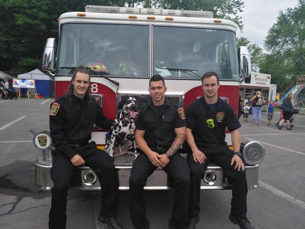 Photograph of three men in firefighters’ office uniforms, sitting on the front of a fire truck with a Dalmatian.