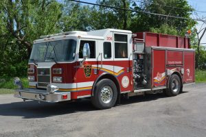 Photograph of a red and white fire truck parked in front of a group of leafy trees.