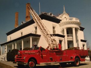 Photograph of a red convertible fire truck raising its aerial ladder in front of a white 19th-century mansion.