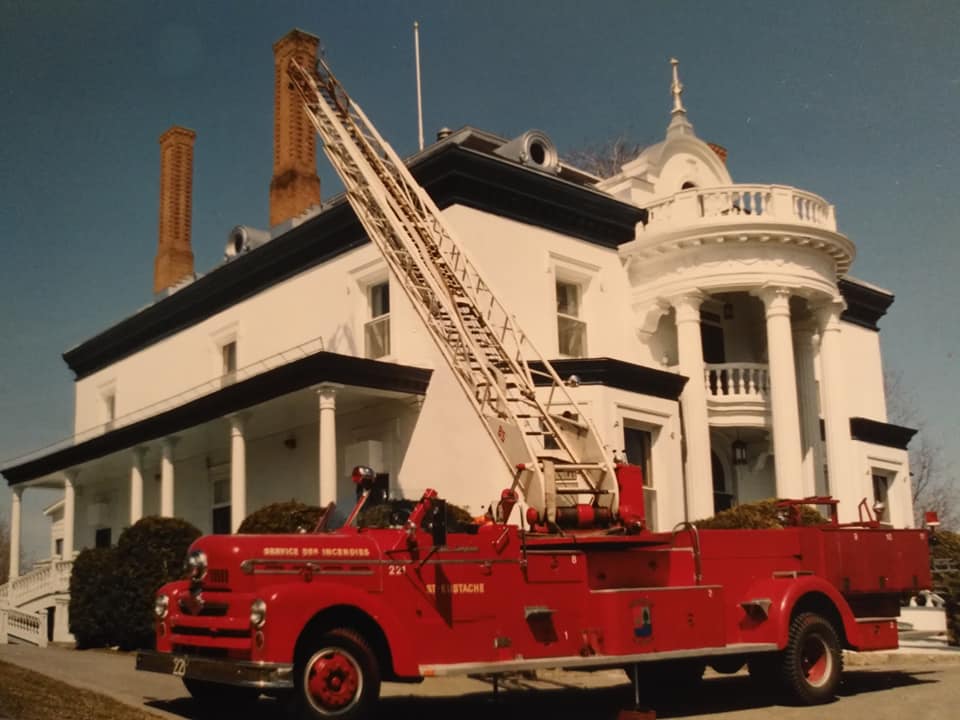 Photograph of a red convertible fire truck raising its aerial ladder in front of a white 19th-century mansion.