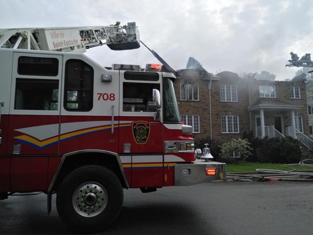 Photograph of a red and white fire truck in front of a row of townhouses on fire. The aerial ladder is pointed toward the location of the fire.