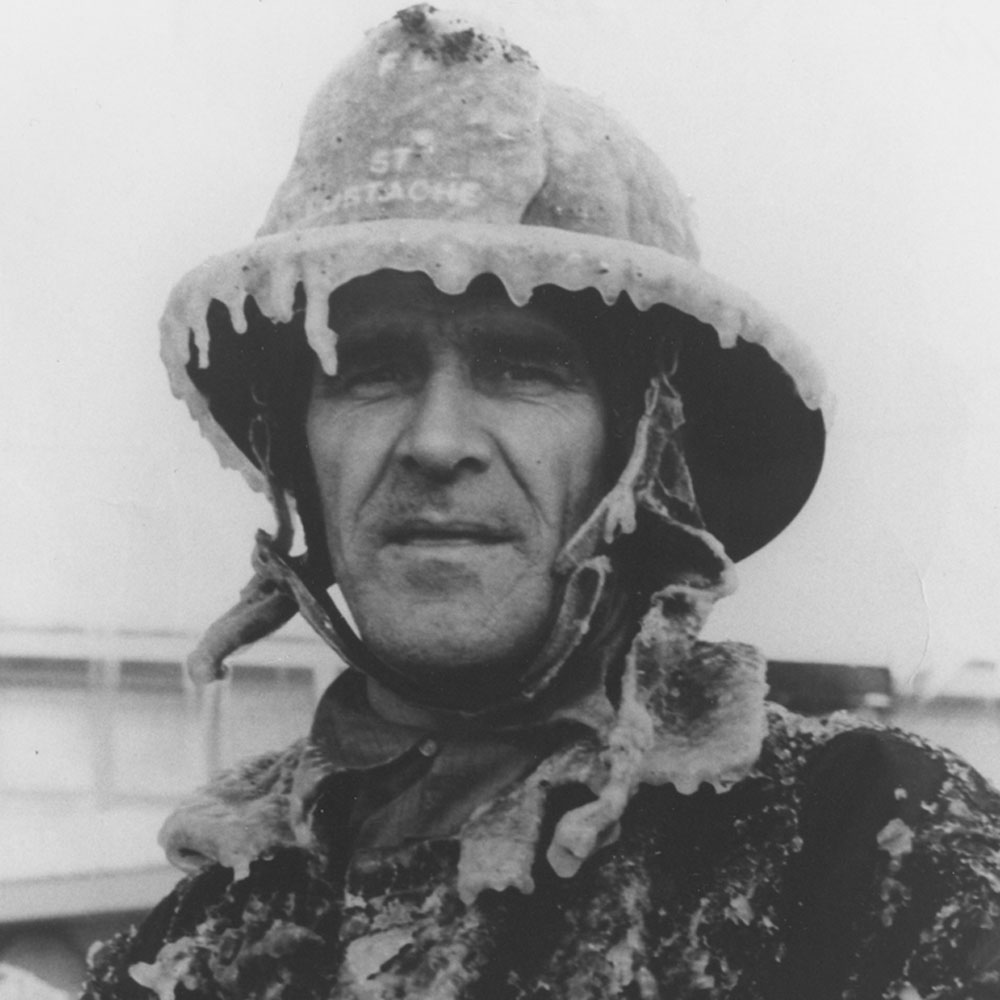 Photographie en noir et blanc d’un homme au visage paisible, arborant un uniforme de pompier couvert de glace. 