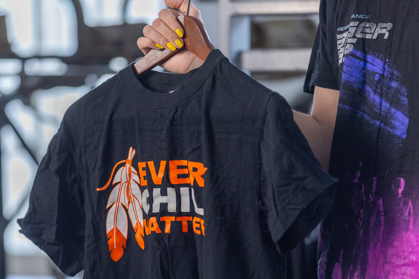 Black shirt on a wooden hanger. It has orange and white text reading EVERY CHILD MATTERS. To the left of the text is a orange and white graphic of two feathers.