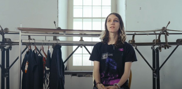 Person sitting in front of a track sewing machine, next to a clothing rack with a few articles of clothing on it.