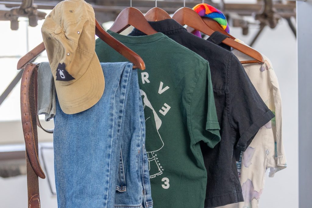 Articles of clothing hanging on five wooden hangers, primarily in neutral colours, with the exception of a rainbow-striped handkerchief tied around the neck of one hanger.