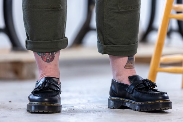 Pair of legs photographed from mid-calf down. The person is wearing black Dr. Marten loafers and cuffed, dark green pants. Multicoloured tattoos are visible on her legs.