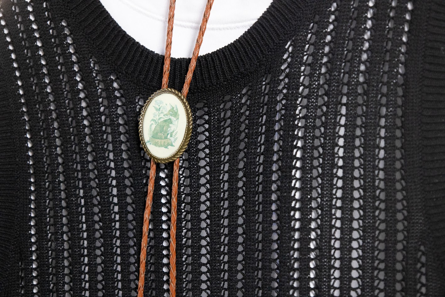 A light brown leather bolo tie, held together by an oval pendant with a green drawing of a fox. The bolo is hanging in front of a black knit top, over a white shirt.