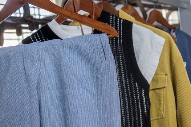 Articles of clothing on wooden hangers, hanging from a rack. The clothes range in colour and style, including blue and white twill pants, a black knit vest, and a yellow corduroy jacket.