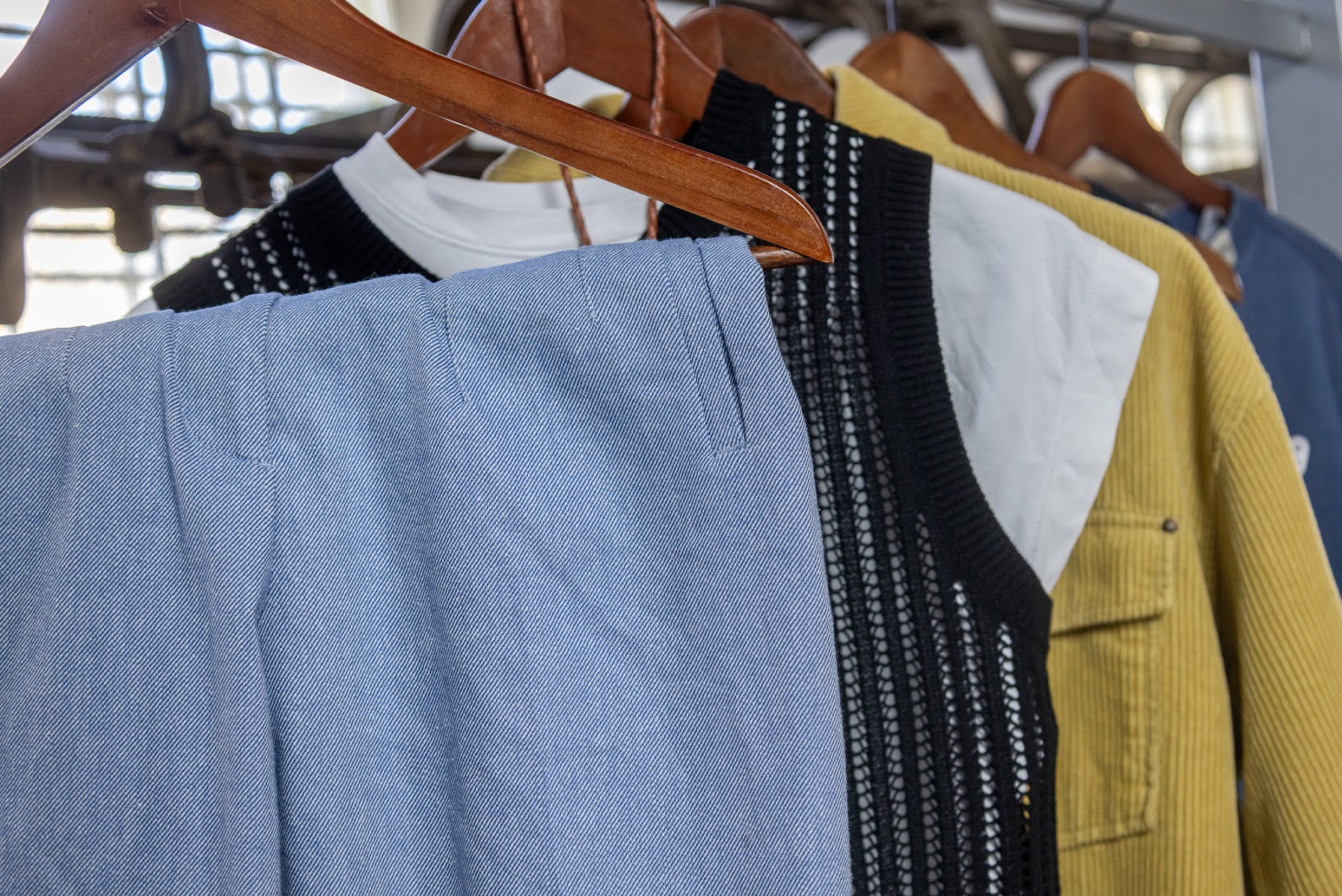 Articles of clothing on wooden hangers, hanging from a rack. The clothes range in colour and style, including blue and white twill pants, a black knit vest, and a yellow corduroy jacket.