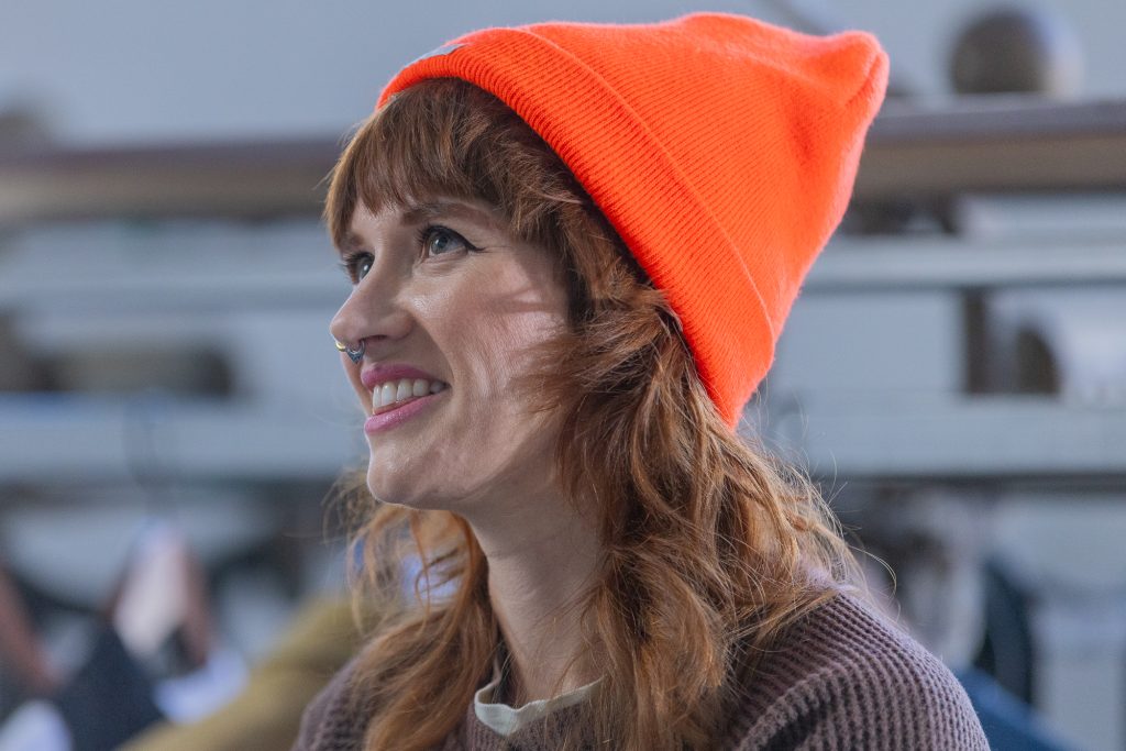 Portrait photo of someone angled to the left, smiling, and looking up. They are wearing a bright orange knit hat.